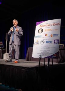 image of man speaking in front of sign with sponsor logos