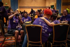 people smiling in matching purple shirts; man taking a picture of others